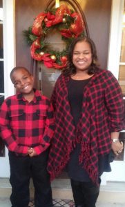Chenita and son wearing coordinating red and black flannel shirts stand in front of a Christmas wreath